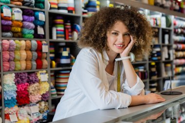 young saleswoman with needle cushion on hand smiling at camera near counter and needlework accessories clipart