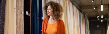 curly woman smiling while looking at multicolored curtains in textile shop, banner clipart