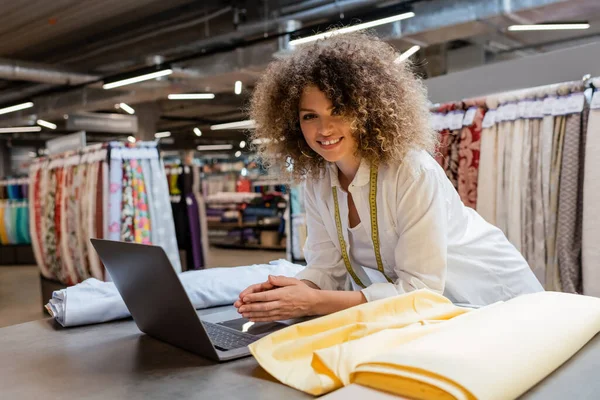 纺织品商店里 一个快乐的卷发女售货员在面料滚筒旁边用笔记本电脑 — 图库照片