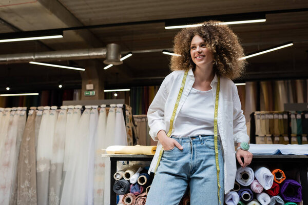 cheerful saleswoman with curly hair standing with hand in pocket near rack with fabric rolls  