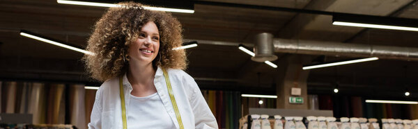 cheerful saleswoman with curly hair standing in textile shop, banner