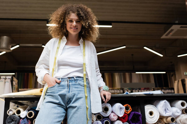 pleased saleswoman with curly hair standing with hand in pocket near rack with different fabric rolls  