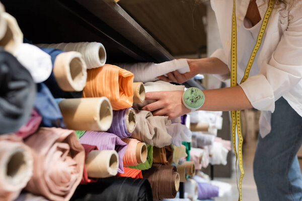 cropped view of saleswoman choosing between colorful fabric rolls on shelves of textile shop