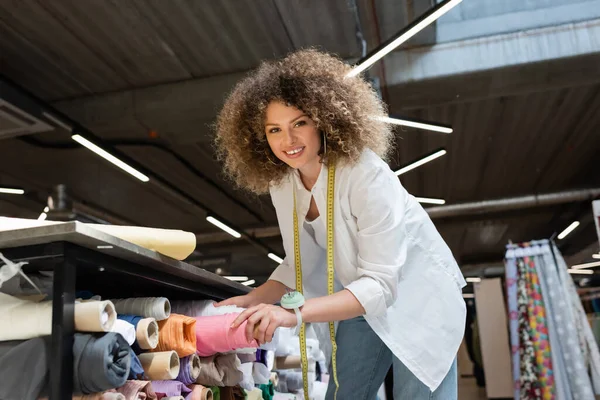 stock image cheerful saleswoman taking pink fabric roll from shelf in textile shop