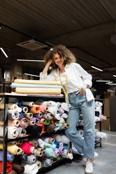 full length of positive saleswoman posing with hand in pocket while leaning on rack with colorful fabric rolls 