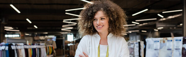 curly saleswoman smiling and looking away near assortment of textile on blurred background, banner