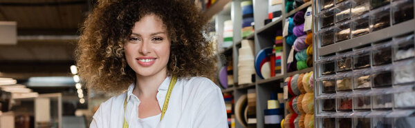 pretty and happy saleswoman looking at camera near rack with needlework accessories in textile shop, banner