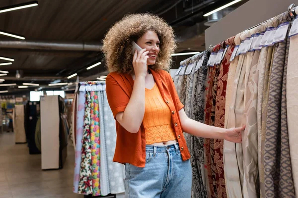 在纺织店里用智能手机笑着卷曲的女人摸布料 — 图库照片
