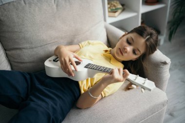 Evdeki kanepede uzanmış küçük Hawai gitarı çalan genç kızın yüksek açılı görüntüsü.