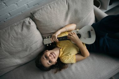 top view of joyful girl lying on sofa and playing small hawaiian guitar clipart