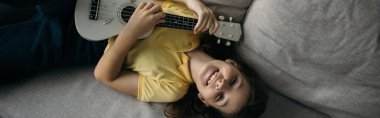 Kanepede uzanan ve kameraya bakarken küçük Hawai gitarı çalan neşeli kız manzarası.