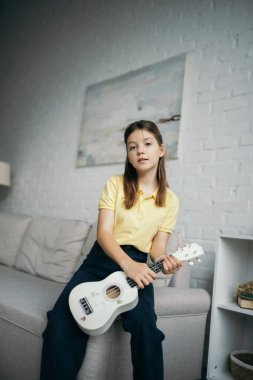 Küçük Hawaii gitarlı genç kız oturma odasındaki kanepenin yanındaki kameraya bakıyor.