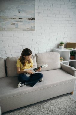 Çapraz bacaklı ve ukulele çalan modern koltukta oturan bir kızın yüksek açılı görüntüsü