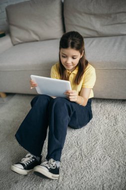 Dijital tabletli gülümseyen kız kanepenin yanında oturuyor.