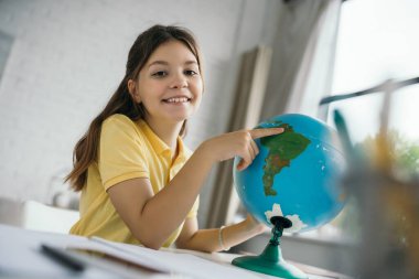 Neşeli kız dünyayı işaret ediyor ve bulanık ön planda evde eğitim alırken kameraya gülümsüyor.