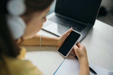 blurred kid holding smartphone with blank screen near laptop at home clipart