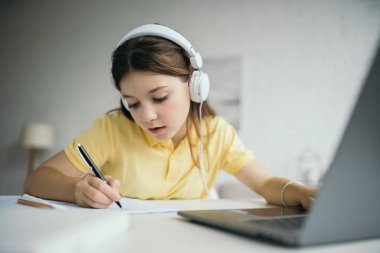 Kablolu kulaklıklı liseli kız bilgisayarın yanında bulanık ön planda not defteri yazıyor.