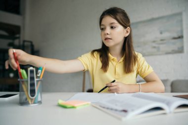 schoolgirl choosing pen near notepad on blurred foreground at home clipart