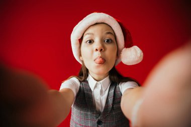 funny girl in santa hat sticking out tongue on blurred foreground isolated on red clipart