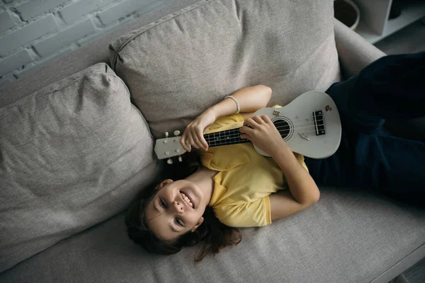 Vista Superior Menina Alegre Deitada Sofá Tocando Pequena Guitarra Havaiana — Fotografia de Stock