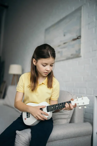 stock image preteen girl sitting on couch in blurred living room and playing ukulele