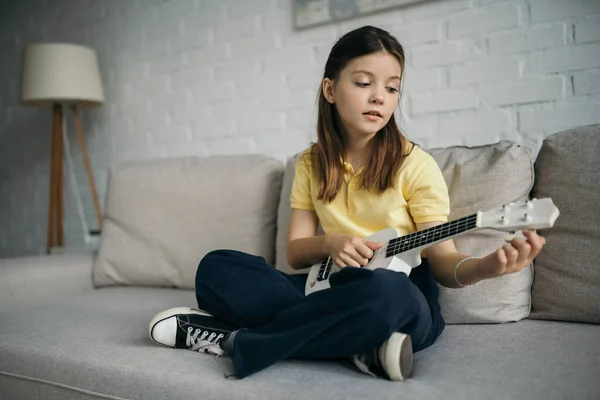 黑发女孩坐在沙发上 双腿交叉 弹奏着小曲的哈瓦伊吉他 — 图库照片