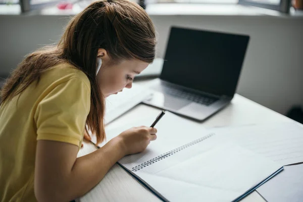 在家里的笔记本电脑旁 一个黑发女孩正在用无线耳机在笔记本上书写 — 图库照片