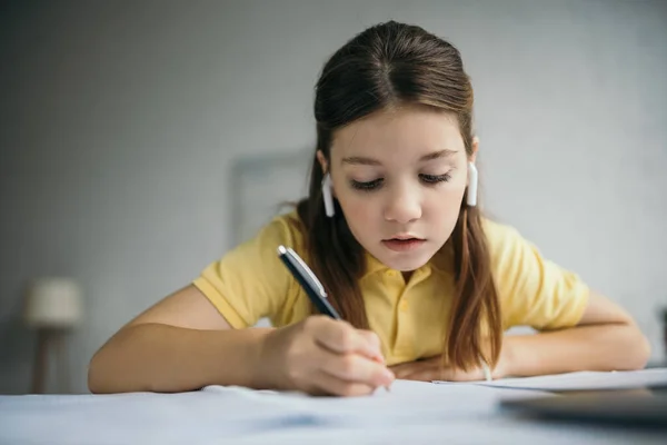 在做家庭作业的同时 用无线耳机在笔记本上写字的少女 — 图库照片