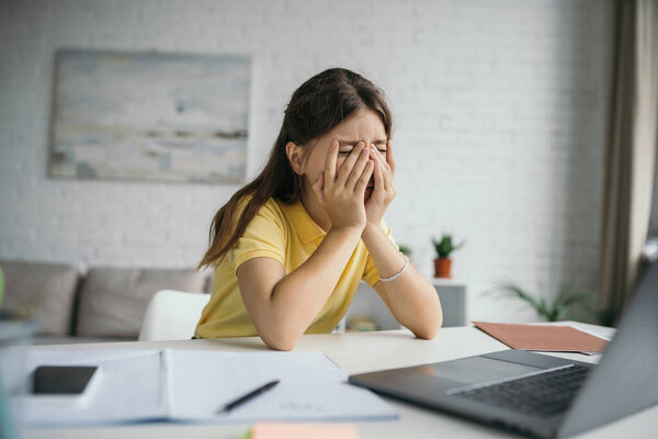 exhausted kid with closed eyes covering face with hands while sitting near blurred laptop at home