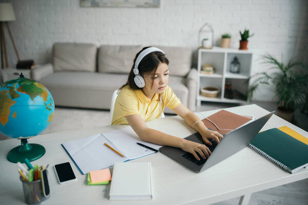 high angle view of girl in headphones typing on laptop near globe and copybooks