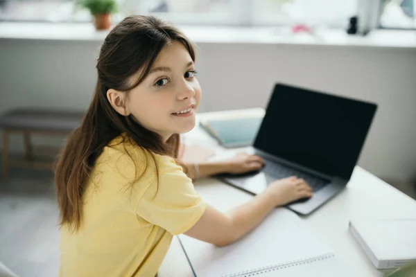 在家里念书的时候 黑发女孩对着相机笑 在模糊的笔记本电脑上打字 — 图库照片