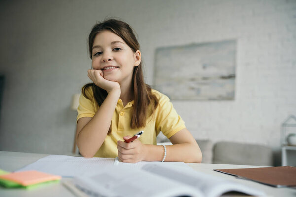 cheerful girl looking at camera while sitting with pens at blurred table