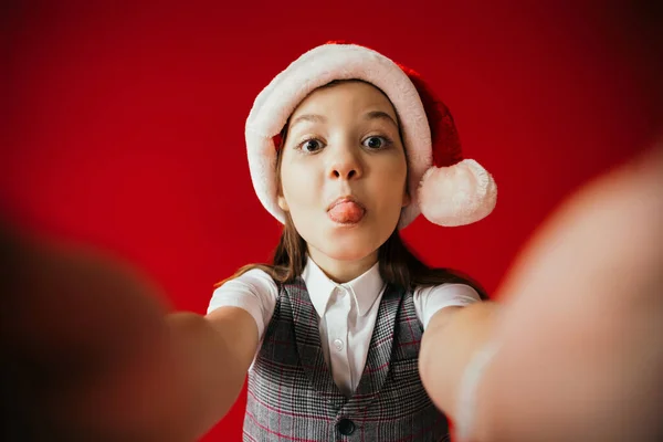 funny girl in santa hat sticking out tongue on blurred foreground isolated on red