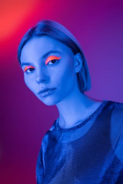 Mor ve mercan arka planında mavi neon ışıkta parlayan makyajlı genç bir kadın.