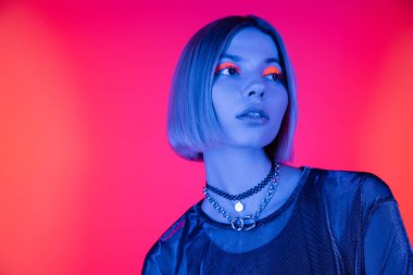Neon makyajlı şık bir kadın mercan pembe arka planda mavi ışıkta uzağa bakıyor.