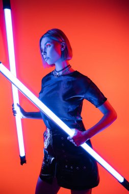 Genç kadın mercan kırmızısı arka planda parlayan neon lambalarla poz verirken başka tarafa bakıyor.