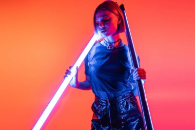 Mercan ve pembe arka planda duran mavi neon lambalı genç ve şık bir kadın.