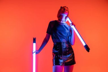 Moda tişörtlü ve deri mini etekli genç bir kadın mercan kırmızısı arka planda parlak neon lambalarla poz veriyor.