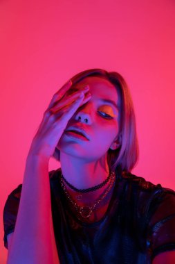 Neon makyajlı ve kolyeli bir kadın. Yüzü silik, eli mor, arka planda pembe bir kadın.