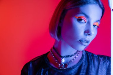 Kolyeli ve parlak neon makyajlı bir kadın mercan pembe arka planda mavi ışıkta kameraya bakıyor.