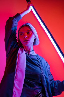 Mercan ve pembe arka planda parlayan neon lambayı tutan ceketli genç bir kadın.