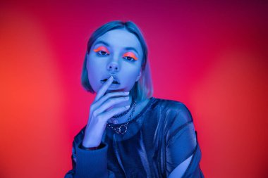 Neon makyajlı genç bir kadın mercan ve pembe arka planda mavi ışıkta dudaklarına dokunuyor.