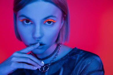 Parlak makyajlı şehvetli bir kadın mercan pembe arka planda mavi neon ışıklı dudaklara dokunuyor.