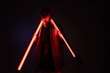 Karanlıktaki genç kadın mor ve siyah arka planda parlayan neon lambalarla ayakta duruyor.