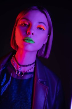 Siyah arka planda mor ışıkta kameraya bakan neon makyajlı ve kolyeli şık bir kadının portresi.