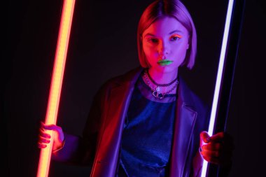 Siyah arka planda mor floresan lambalar tutan neon makyajlı ve deri ceketli güzel bir kadın.