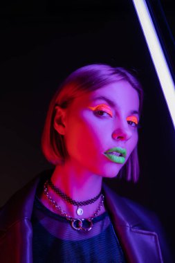 Parlak neon makyajlı modacı kadın siyah arka planda mor floresan lambanın yanındaki kameraya bakıyor.