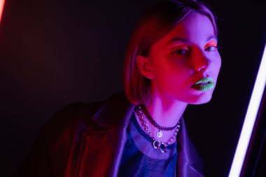 Siyah arka plandaki floresan lambanın mor ışığında kameraya bakan neon makyajlı genç bir kadın.