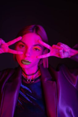 Neon makyajlı ve kolyeli moda bir kadın zafer işaretleri gösteriyor ve siyah arka planda mor ışıkta kameraya bakıyor.