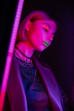 Düşük açılı, parlak makyajlı, koyu arkaplanda mor neon ışığı olan bir kadın.